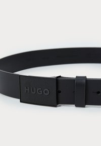 Czarny gumowy pasek z fakturowaną prostokątną klamrą z wytłoczonym napisem "HUGO". Projekt jest minimalistyczny, z wieloma otworami do regulacji.