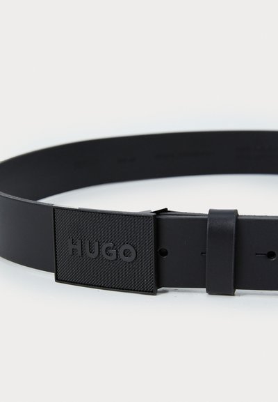 Ceinture en caoutchouc noir avec une boucle rectangulaire texturée portant l'inscription "HUGO" en relief. Le design est minimaliste avec plusieurs trous d'ajustement.