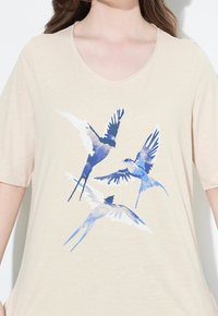 Camisa beige con un gráfico en estilo acuarela en azul y blanco de tres golondrinas en vuelo en el pecho.