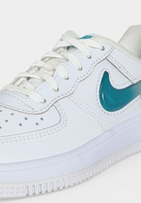 Λευκό αθλητικό παπούτσι Nike με τυρκουάζ σήμα, λευκά κορδόνια και διάτρητη λεπτομέρεια στη μύτη, σε απλό ανοιχτό φόντο.