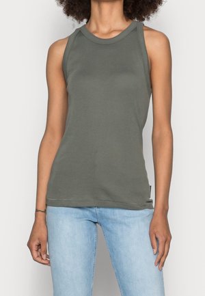 Woman wearing sleeveless olive green tank top and light blue jeans, standing against a plain light background.