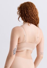 Eine rothaarige Person mit Sommersprossen, die eine beige BH und Unterwäsche trägt. Sie zeigt Tätowierungen am linken Arm und am Nackenrücken vor einem hellen Hintergrund.