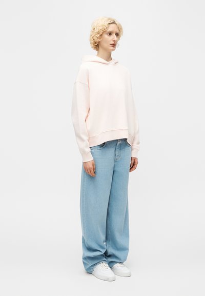 Άτομο με κοντά σγουρά ξανθά μαλλιά που φοράει ανοιχτό ροζ φούτερ με κουκούλα, φαρδιά ανοιχτά μπλε τζιν και λευκά αθλητικά παπούτσια, στέκεται μπροστά σε απλό λευκό φόντο.