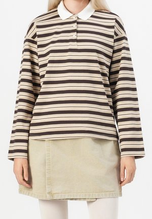 Frau trägt ein langärmeliges, braun-beige gestreiftes Polohemd mit weißem Kragen, einen beigen Rock und weiße Leggings.