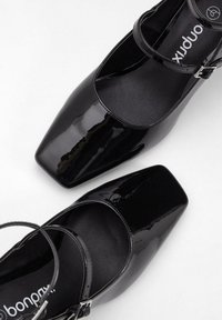 Scarpe in vernice nera con punta quadrata, cinturino alla caviglia e design minimalista. Texture liscia e finitura elegante. Nome del marchio visibile all'interno.
