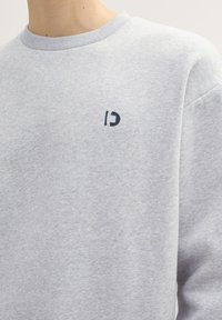 Sivá mikina vyrobená z mäkkej tkaniny, s okrúhlym výstrihom a malým námorníckym logom na hrudi. Pohodlný strih a jednoduchý dizajn.