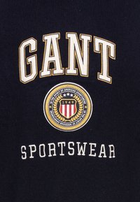Felpa blu navy con ricamo bianco e giallo "GANT", dotata di un logo circolare e del testo "SPORTSWEAR" sottostante.