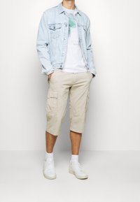 Chaqueta de mezclilla azul claro, camiseta gráfica blanca, pantalones cortos beige tipo cargo y zapatillas blancas con calcetas, de pie contra un fondo claro.