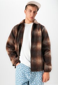 Jonge man in een bruin en zwart verloop ritsjack, wit T-shirt, lichtblauwe broek met stippen en een witte pet poseert tegen een witte achtergrond.