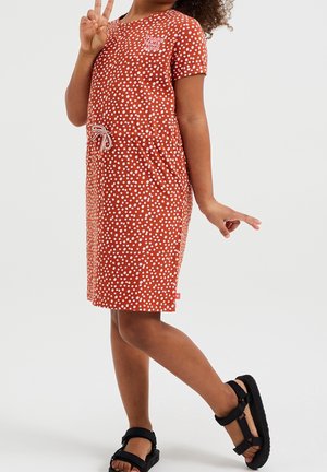 Ung person iført en rød polkaprikket kjole og sorte sandaler, poserende med det ene ben løftet og hånden viser tre fingre.