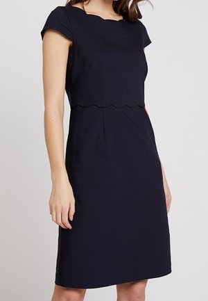 Femme portant une robe noire sans manches avec un détail festonné à la taille sur un fond clair uni.