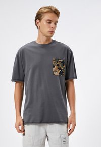 Camiseta gris de manga corta con cuello redondo y un bolsillo en parche de camuflaje en el lado izquierdo del pecho, hecha de un suave tejido de algodón.