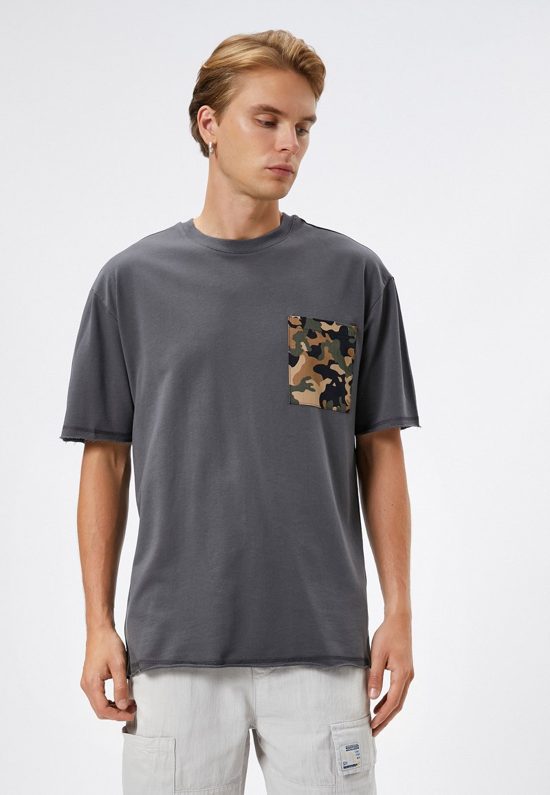 Camiseta gris de manga corta con cuello redondo y un bolsillo en parche de camuflaje en el lado izquierdo del pecho, hecha de un suave tejido de algodón.