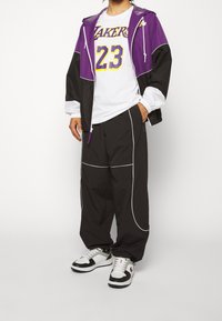 Chaqueta de deporte púrpura y negra, camiseta blanca con "Lakers 23", pantalones negros con ribete blanco y zapatillas negras y blancas.
