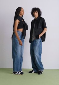 To personer poserer mot en lys bakgrunn. Den første har på seg en svart topp, høyt liv med jeans og svarte joggesko. Den andre har en svart skjorte over en grå topp, kombinert med vide blå jeans og svarte sko.