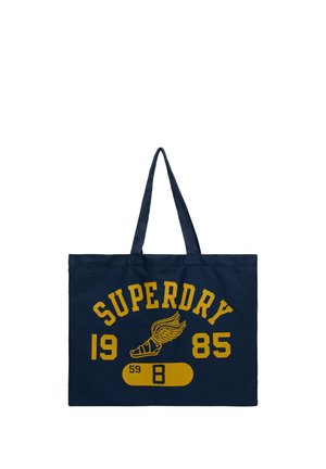 Mørkeblå totebag med gul tekst "SUPERDRY 1985" og en vinget sandal-grafik over et gult pilleformet design med tal.