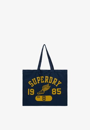 Mørkeblå totebag med gul tekst "SUPERDRY 1985" og en vinget sandal-grafik over et gult pilleformet design med tal.