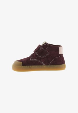 Scarpa da bambino in pelle scamosciata viola scuro con strap a velcro, toppa beige sul tallone, cuciture bianche e suola in gomma marrone traslucida.