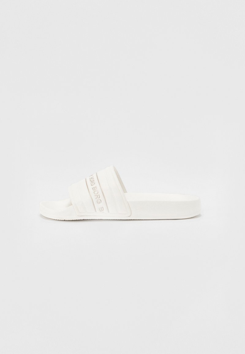 Vit slidesandal med en strukturerad rem, som har horisontella riller och präglad logotyp. Platt sula med en slät yta.