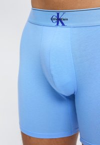 Μπλε ανδρικά μπόξερ Calvin Klein, φορεμένα στη μέση, με λογότυπο στη μέση της ζώνης και υφή ελαστικού, εφαρμοστού υφάσματος.