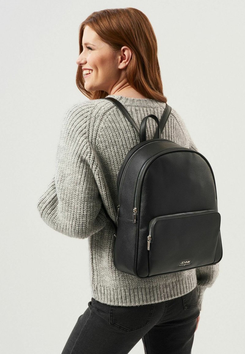 Czarny skórzany plecak o gładkiej teksturze, z podwójnymi zamkami błyskawicznymi, oraz przednią kieszenią. Regulowane paski i zaokrąglony design u góry.