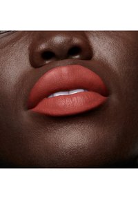 Rouge à lèvres en terre cuite mate appliqué sur des lèvres pleines et lisses, contrastant avec une peau brun profond, mettant en valeur une texture subtile et un éclat naturel.