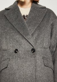 Primer plano de una persona que lleva un abrigo gris de lana de doble botonadura con solapas anchas y botones negros, sobre un suéter beige con patrones.