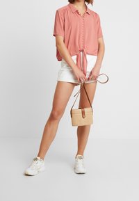 Chemise courte à manches courtes, coupée, en corail doux avec un nœud à l'avant. Associée à un short blanc et un petit sac tissé beige. Baskets blanches.