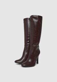 Bottes montantes bordeaux en cuir brillant. Présentent un talon fin et une sangle avec une boucle en argent accent sur la cheville.