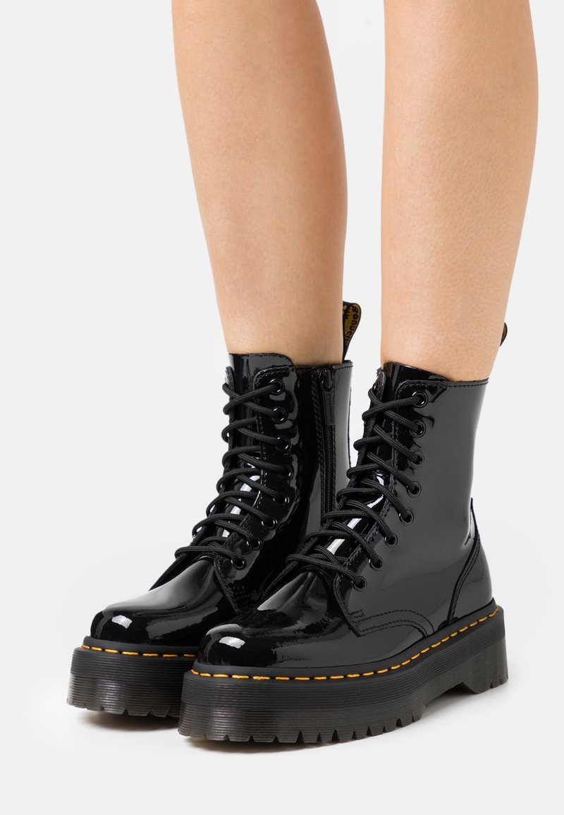 Bottes en cuir verni noir brillant à lacets, semelles épaisses et coutures jaunes, portées sur des jambes nues sur un fond uni.