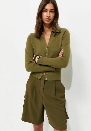 Vrouw met bruin haar die een olijfgroene, dichtgeknoopte cardigan draagt en bijpassende cargoshorts tot op de knie, staand met de armen over elkaar.