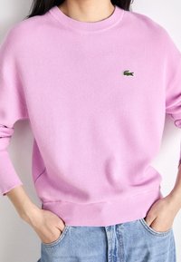Sweat-shirt en maille rose clair avec col rond et poignets côtelés. Présente un petit logo Lacoste vert sur la poitrine. Porté sur un jean bleu.
