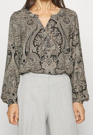 Mujer con blusa de manga larga con estampado de paisley, metida dentro de pantalones de talle alto de color gris claro, de pie contra un fondo liso.