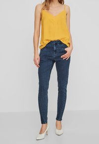 Top amarillo sin mangas con escote en V, combinado con jeans ajustados de color azul oscuro. La modelo lleva zapatos de tacón alto blancos. Diseño y textura simples.