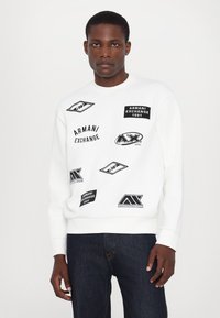 Biała bluza z różnymi czarnymi graficznymi naszywkami, w tym logo marek i tekstem. Klasyczny okrągły dekolt i ściągacze. Casualowy design.