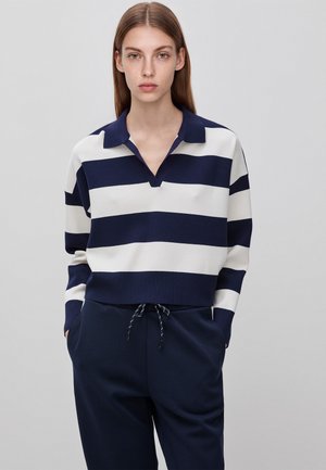 Jonge vrouw met lang bruin haar, gekleed in een marineblauw-wit brede gestreepte lange mouw polo en marineblauwe broek, met handen in de zakken.