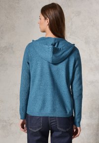 Hoodie en tricot bleu avec un design texturé et une capuche à cordon. Présente des poignets et un ourlet côtelés, associé à un jean en denim foncé.