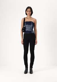 Top bustier din denim, design patchwork în nuanțe de albastru închis, asortat cu blugi negri, slim-fit și cizme cu gleznă, ținând o geantă neagră.