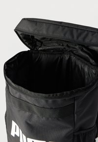 Μαύρο σακίδιο πλάτης Puma με ανοιχτό το κύριο διαμέρισμα και μερικώς ορατό το επάνω φερμουάρ σε απλό φόντο.