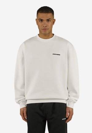 Mann trägt ein weißes Sweatshirt mit dem Text "PEGADOR" auf der Brust und schwarze Hose mit passendem Logo am Oberschenkel, steht aufrecht.