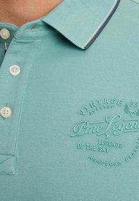 Ljusblå polo med en texturerad tyg, en tre-knapps kn�äppning och en präglad logotypdesign på bröstet. Kontrasterande krage.