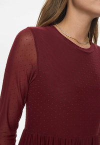 Rochie burgundy cu mâneci lungi din material transparent, având o textură punctată. Fustă evazată și decolteu rotund. Accentuata cu bijuterii delicate din aur.