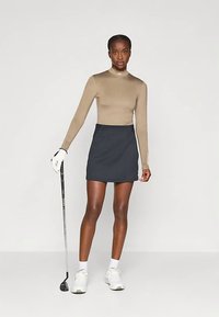 Donna in maglietta a maniche lunghe beige, gonna blu navy, guanto da golf bianco, che tiene un bastone da golf, indossando sneakers bianche su sfondo neutro.