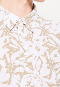 Camicia bianca con un motivo floreale in tonalità beige; presenta un colletto e chiusura con bottoni. Tessuto testurizzato visibile.