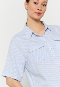 Camicetta corta azzurra in cotone con righe bianche. Presenta una chiusura con bottoni davanti e due tasche frontali.