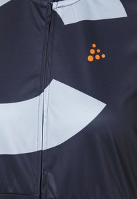 Námornícky a světle modrý látkový materiál s výraznými geometrickými vzory, obsahující vertikální zip a oranžový detail loga. Hladká textura, atletický design.