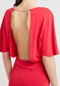 Widok z tyłu osoby w czerwonej sukience z głębokim dekoltem i cienkim złotym łańcuszkiem zapiętym na szyi.