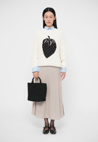 Žena v bielom svetri s veľkým čiernym motívom jahody, béžovou sukňou, čiernymi topánkami, držiaca čiernu kabelku na jednoduchom pozadí.