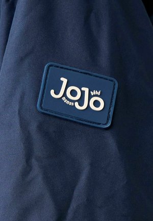 Mørkeblå stofpose med en tekstureret, rektangulær mærkat i blåt med hvid prægede tekst "JoJo maman bébé." Glat materiale, minimalistisk design.
