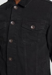 Chaqueta de mezclilla negra con cuello, seis botones frontales, dos bolsillos en el pecho con solapas y detalles de costura sutiles.